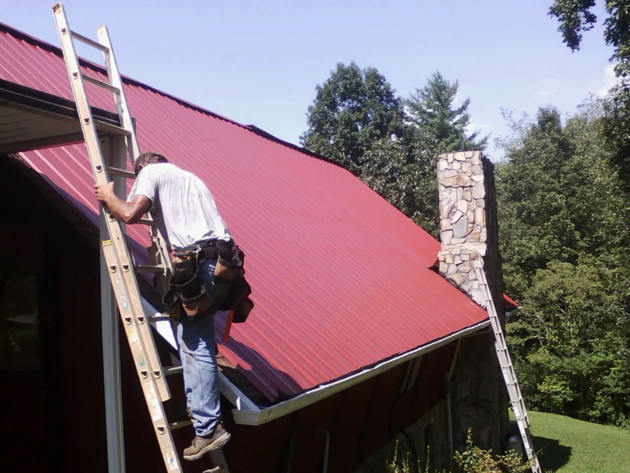 Roofer on a ladder beside a red metal roof and stone chimney — Soffit & Fascia Repair by Roofing Brothers in Osawatomie